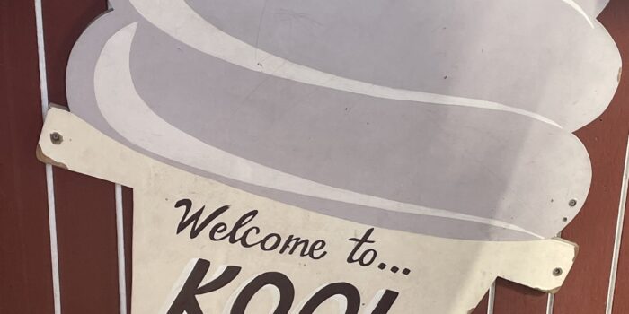 image00006 Welcome to Kool Kone sign in brown lettering on cone of ice cream with soft serve ice cream in cone. Sign located on red siding of building.