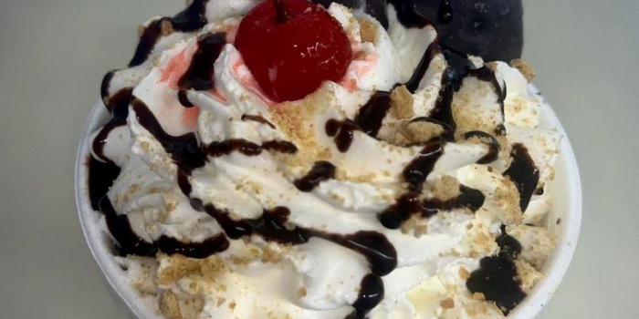 rd26-Kool-Kone-dessert-2023-02-4 Ice cream sundae with a chocolate covered cookie, whipped cream, chocolate sauce drizzle and a red cherry on top.